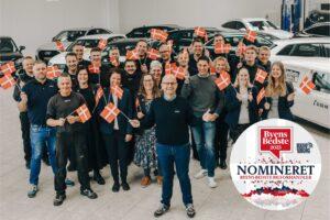Medarbejdere samlet hos Lindholm Biler i biludstilling med flag i hånden og biler i baggrunden i forbindelse med nominering til Byens Bedste Bilforhandler 2023